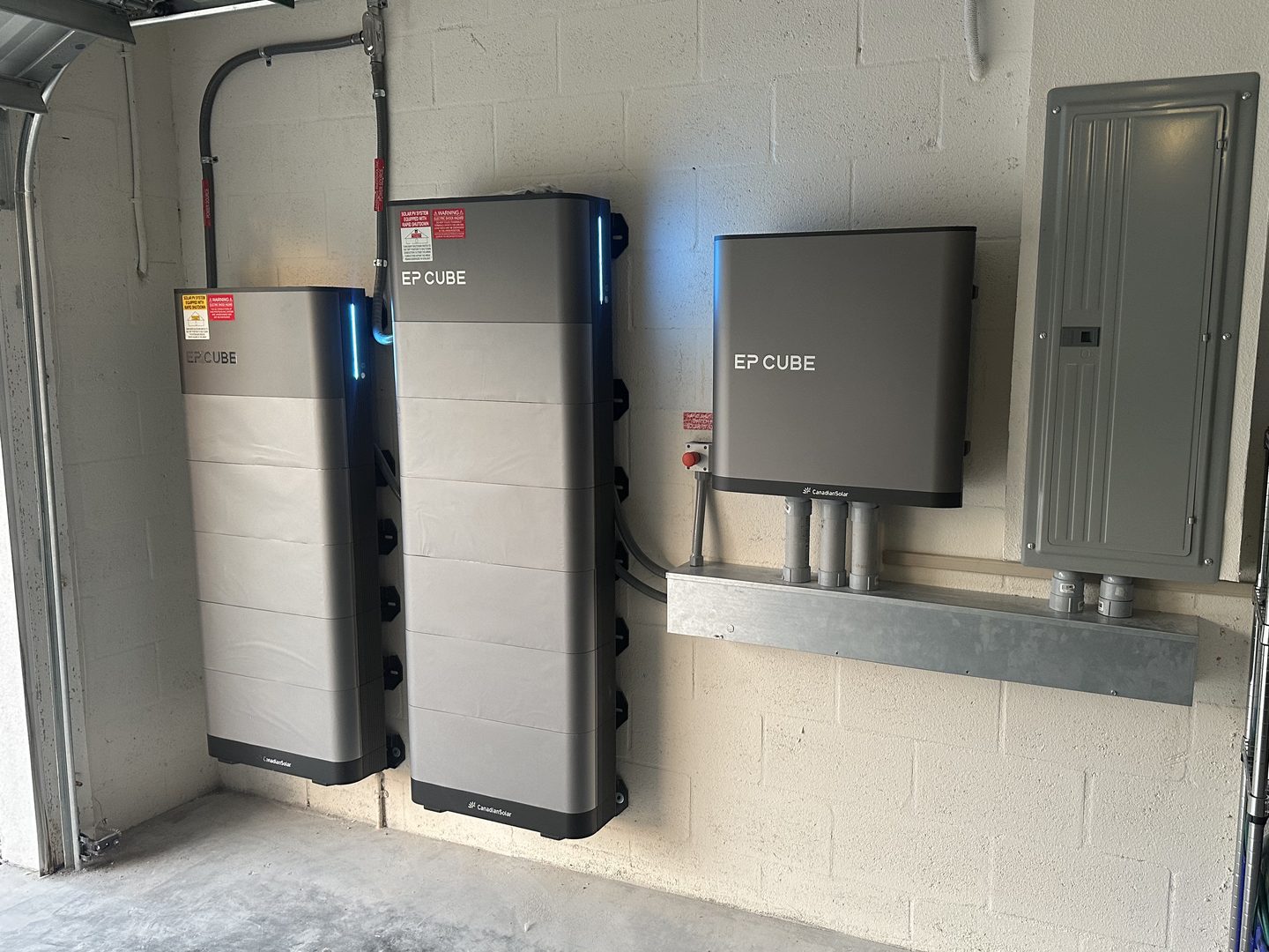 Two battery stacks and a battery inverter mounted in a garage next to the main electrical panel, installed by Ori Energy